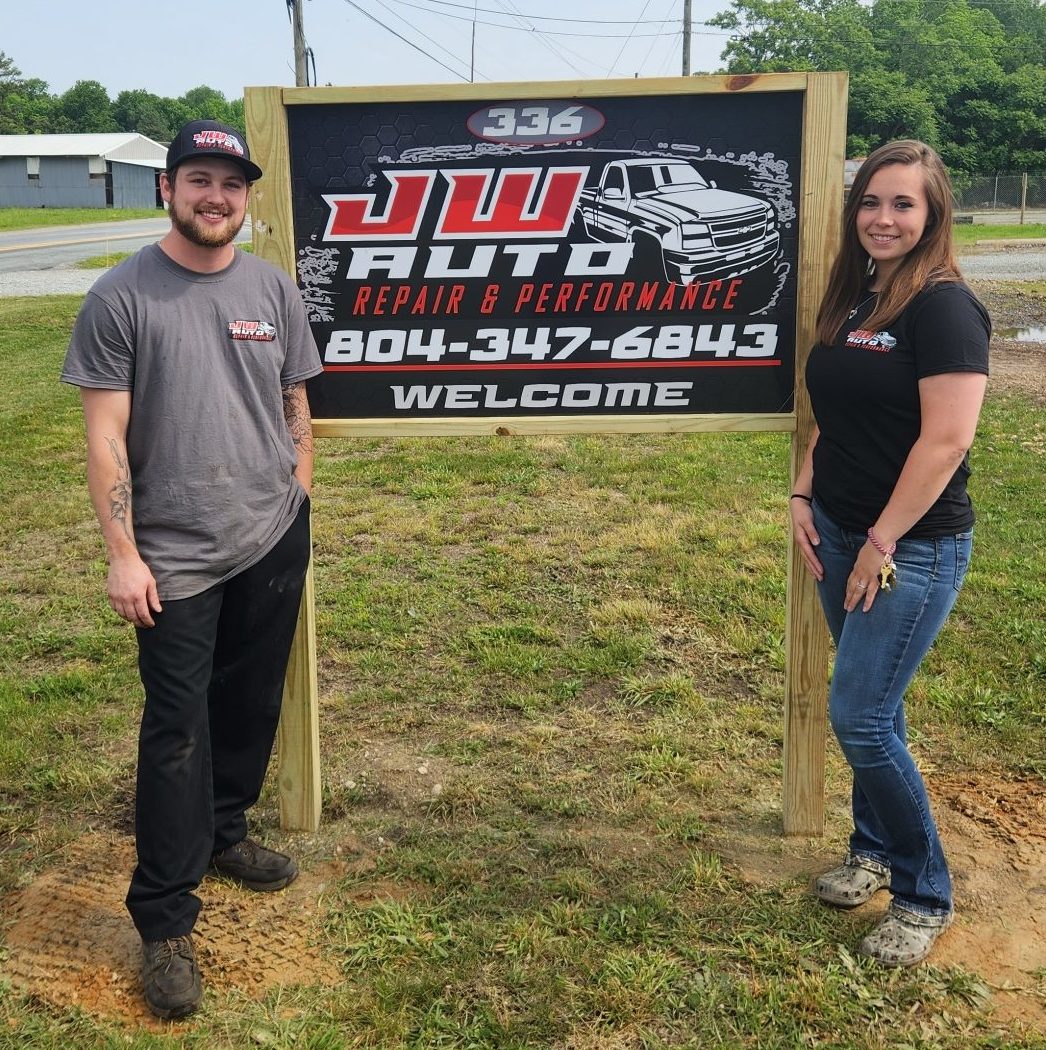 Joey and Kandice standing in front of their business sign in the grass outside of their auto repair shop. The sign reads "JW AUTO REPAIR AND PERFORMANCE 804-347-6843"